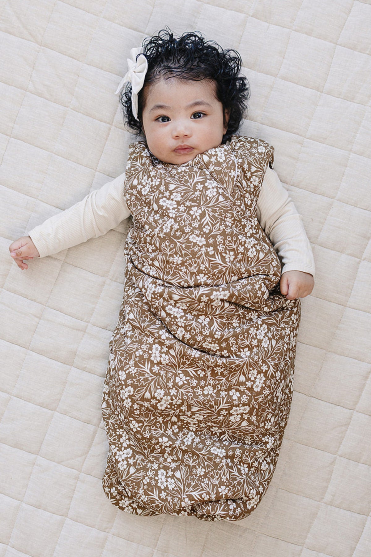A baby with curly black hair lies on a beige quilted mat, wearing a brown floral sleep sack.