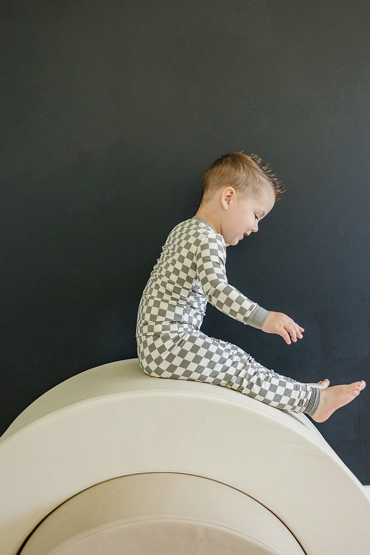 A child in a light green checkered pajama set sits on a beige curved cushion against a dark wall.