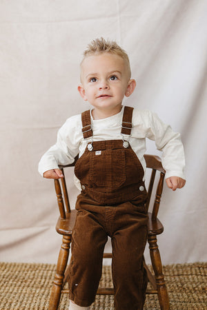 A young child with short, spiky hair sits on a wooden chair, wearing brown corduroy overalls and a white long-sleeve shirt, set against a neutral backdrop.