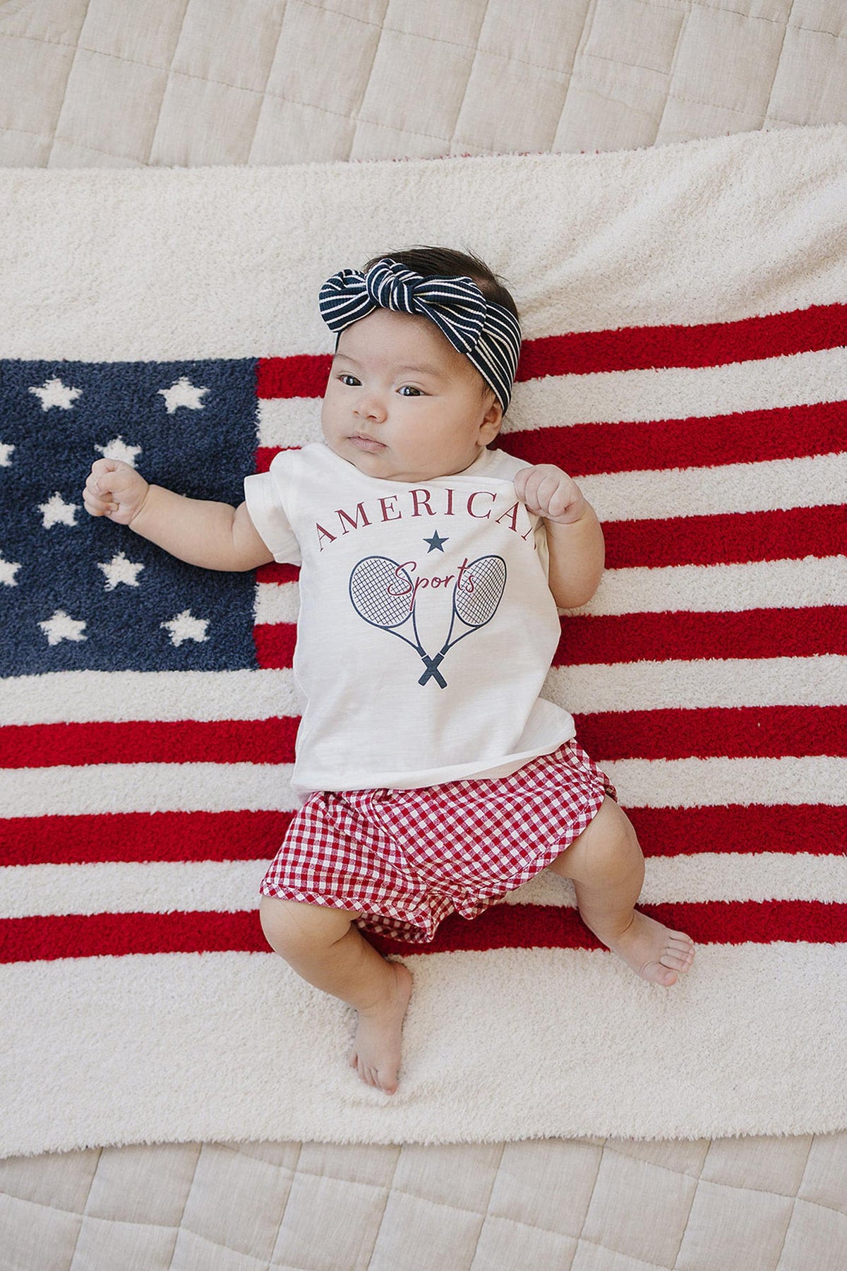 A baby wearing a striped headband and an America Sports shirt lies on a flag-themed blanket.