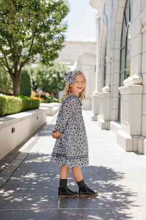 A smiling girl in a floral dress and matching bow stands on a sunny stone pathway beside a building.