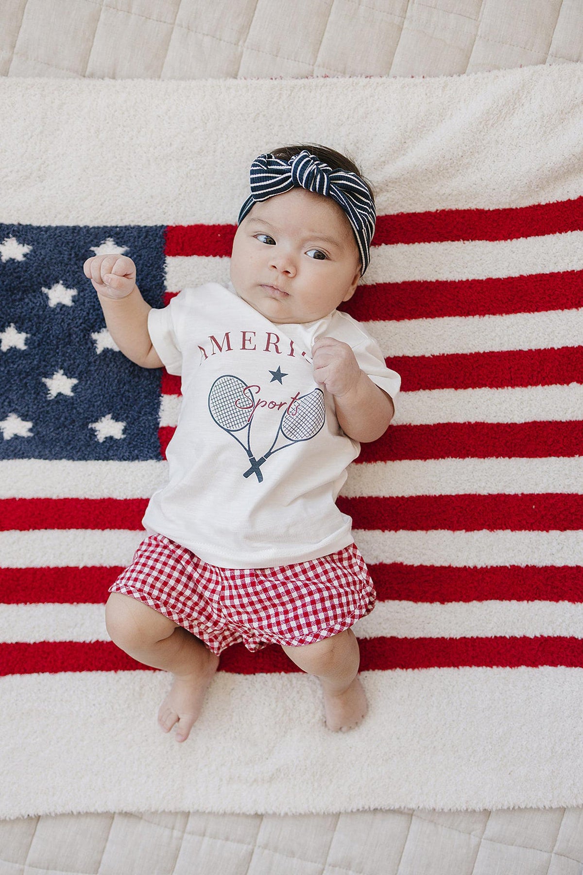 A baby wearing a navy striped head wrap and patriotic outfit lies on an American flag-themed blanket.