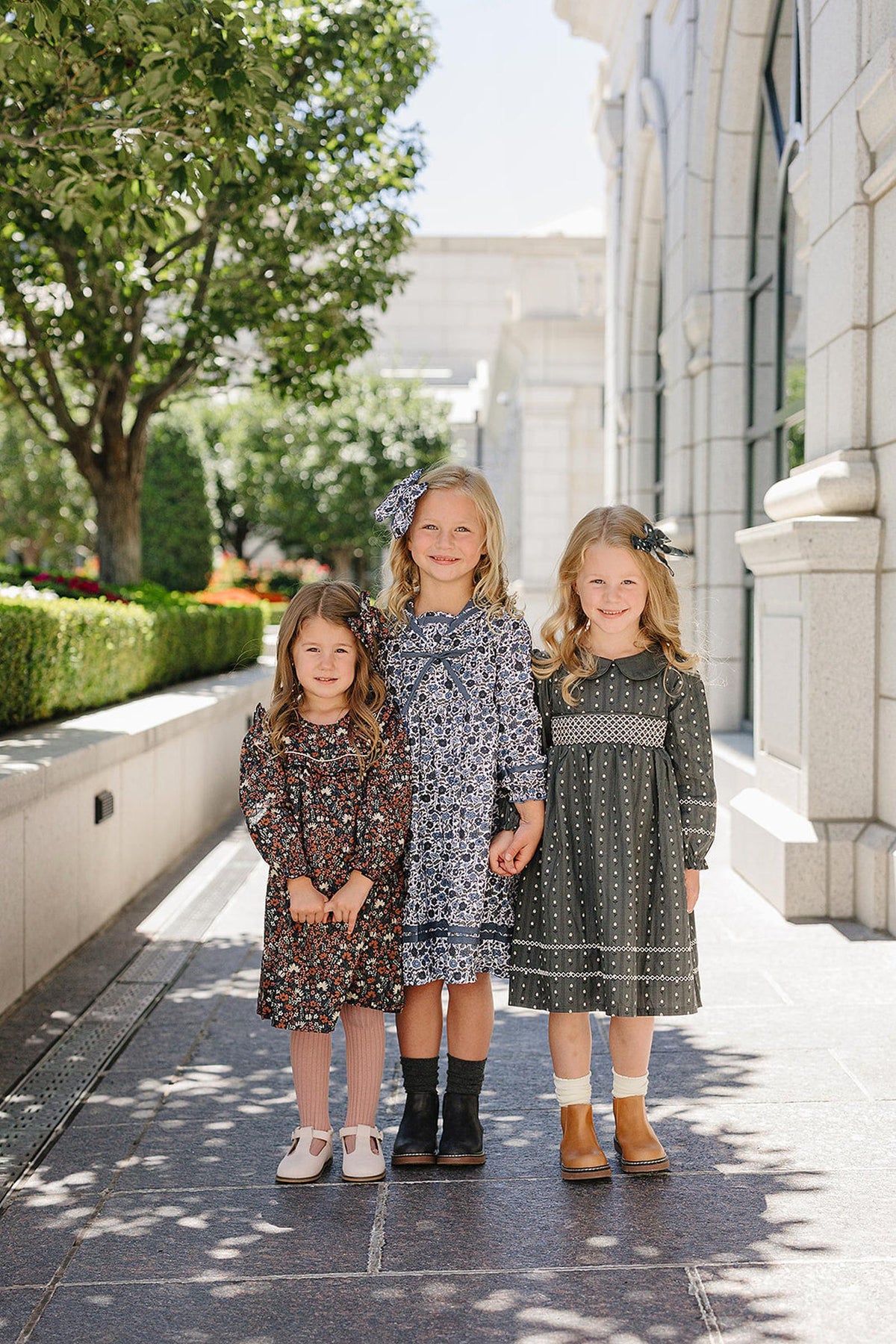 Three young girls in floral dresses stand on a sunlit stone pathway, surrounded by greenery and elegant architecture.