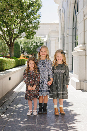 Three young girls in floral dresses stand on a sunlit stone pathway, surrounded by greenery and elegant architecture.
