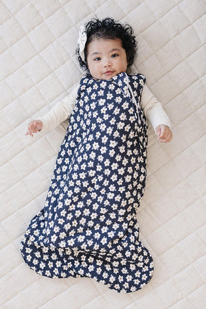 A baby lies on a light-colored quilt, wrapped in a navy sleep sack adorned with white daisy patterns. The soft, breathable fabric ensures comfort for little ones.