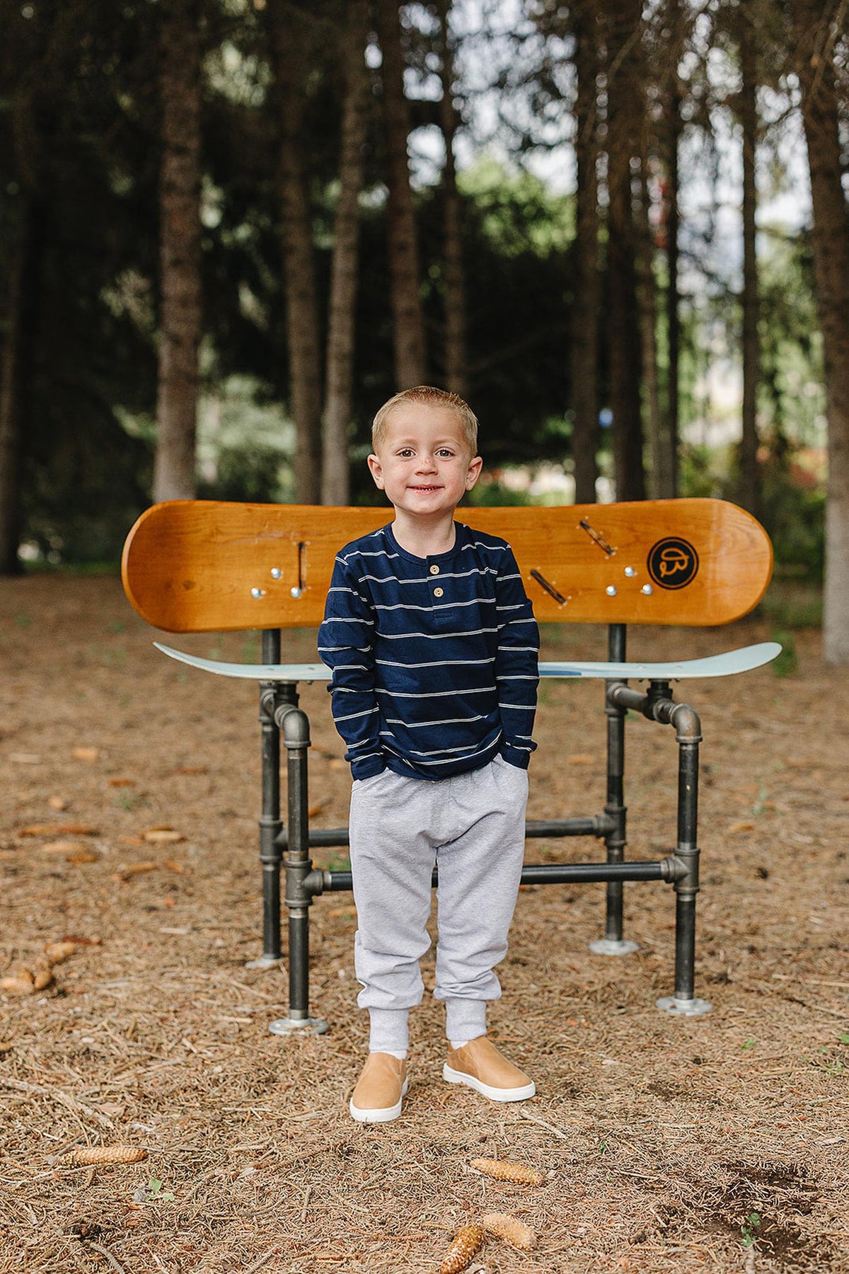 A young boy stands confidently in a forest setting, wearing a navy striped long sleeve shirt and gray sweatpants. He smiles while posing in front of a wooden bench.