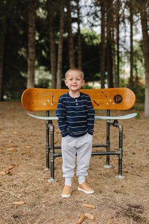 A young boy stands confidently in a forest setting, wearing a navy striped long sleeve shirt and gray sweatpants. He smiles while posing in front of a wooden bench.