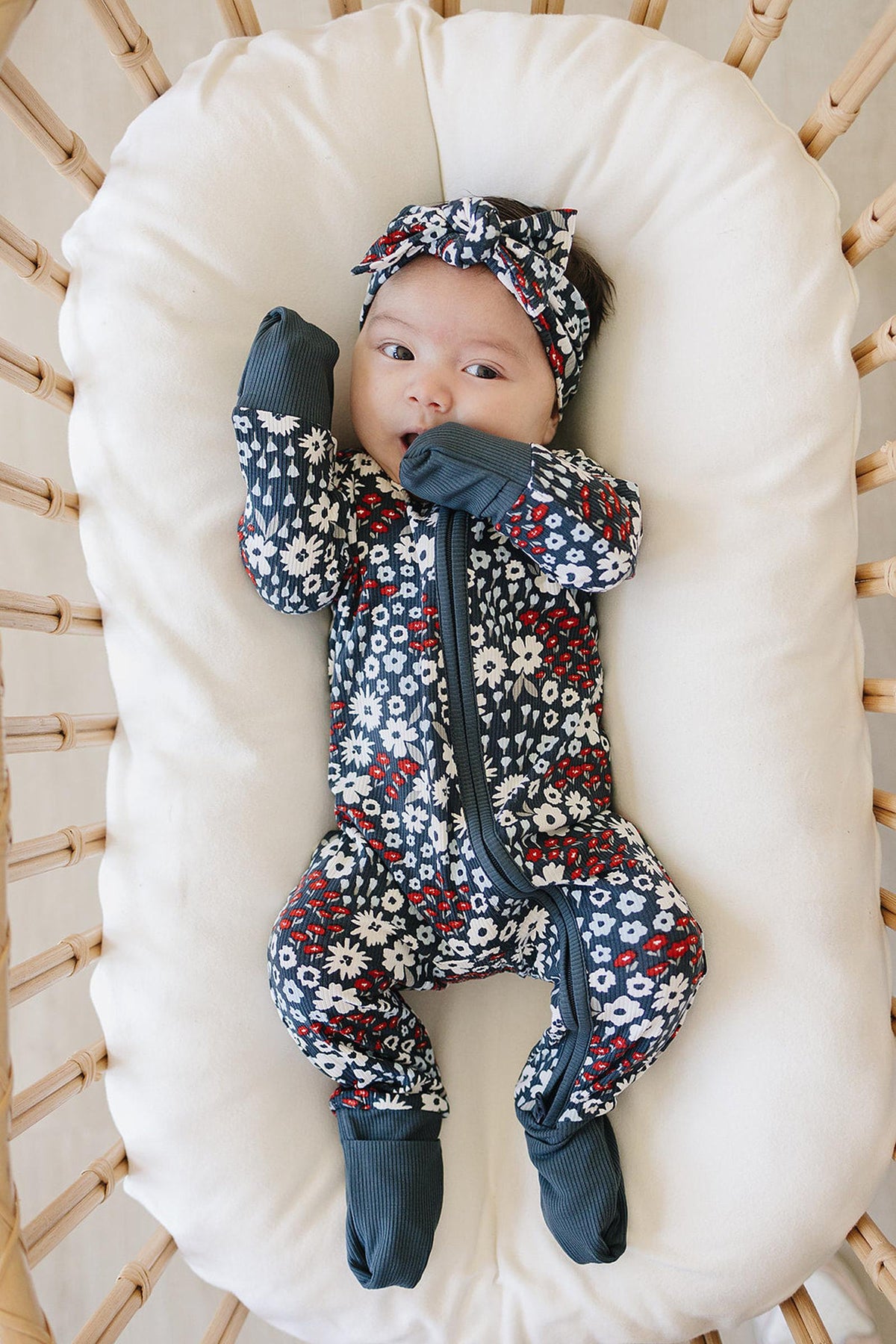 Baby wearing a floral-patterned onesie and matching head wrap, lying on a cushioned wicker chair.