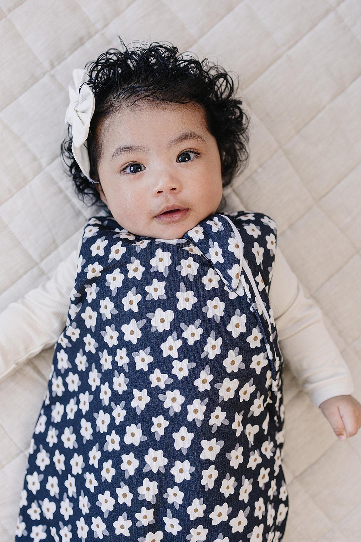 A baby with curly hair is lying on a beige quilt, wearing a navy sleep sack adorned with white daisies. The sleep sack is soft and cozy, perfect for comfort.
