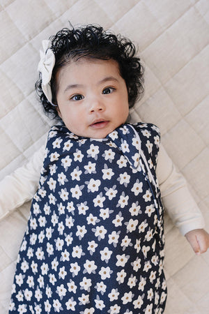 A baby with curly hair is lying on a beige quilt, wearing a navy sleep sack adorned with white daisies. The sleep sack is soft and cozy, perfect for comfort.