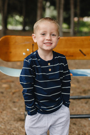 A smiling young boy wears a navy striped long sleeve t-shirt with a two-button placket, paired with light gray pants.