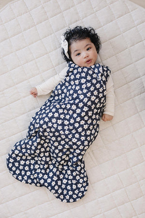 A baby lies on a light quilt, wearing a navy sleep sack adorned with white daisies. The soft fabric is breathable and cozy, perfect for comfort during sleep.