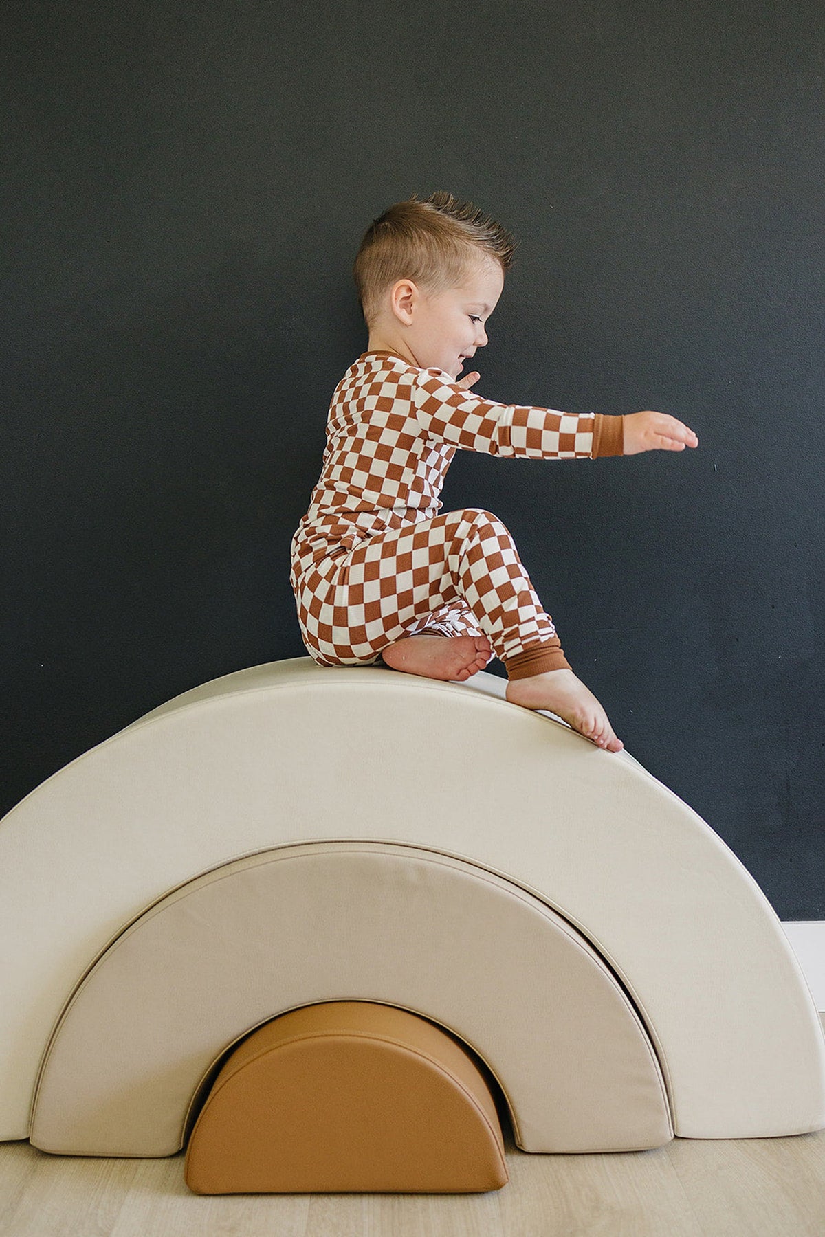 A child in rust checkered pajamas sits on a beige cushioned arch against a dark wall.