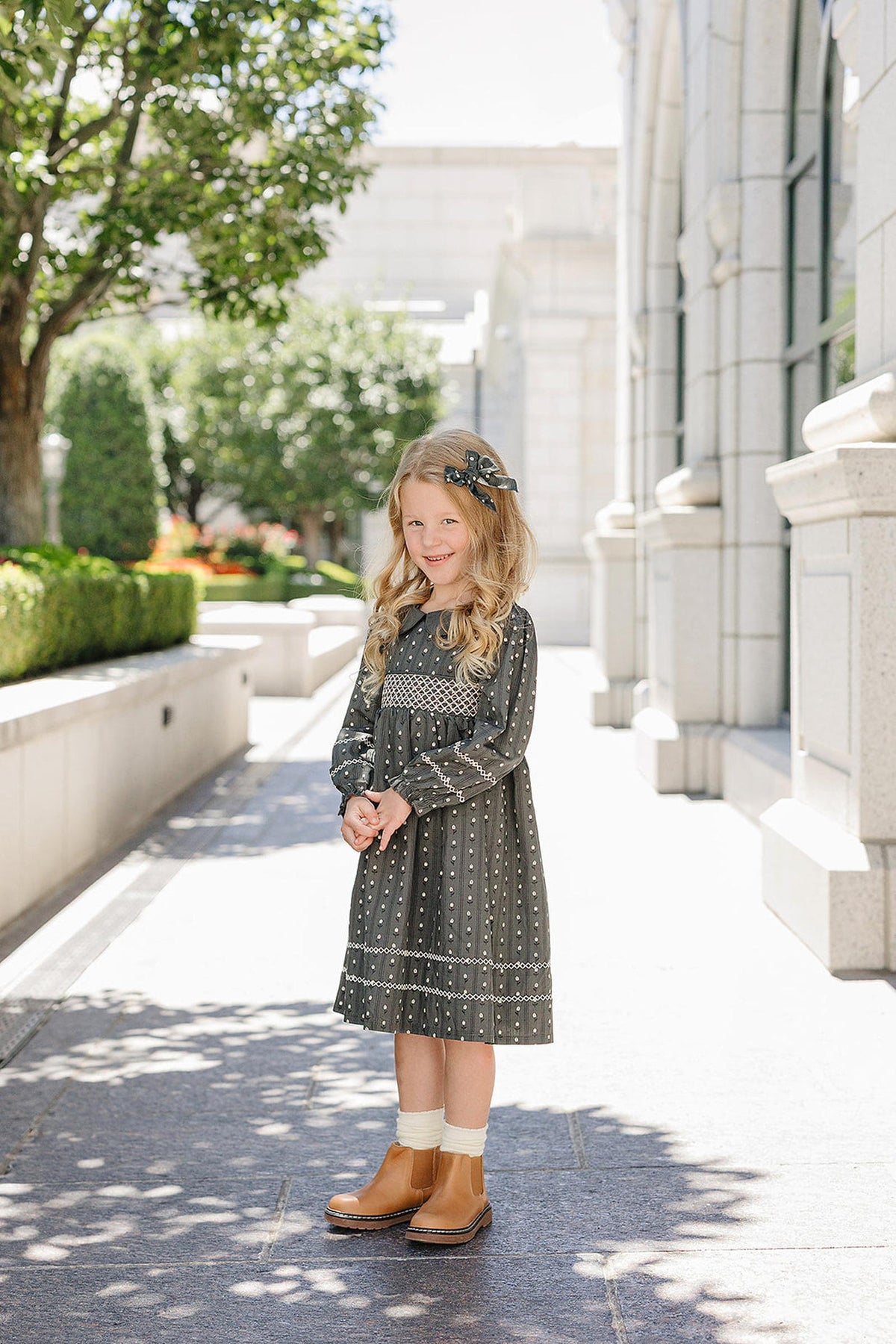 A young girl in a patterned dress and brown boots stands outdoors, wearing a cute hair bow.