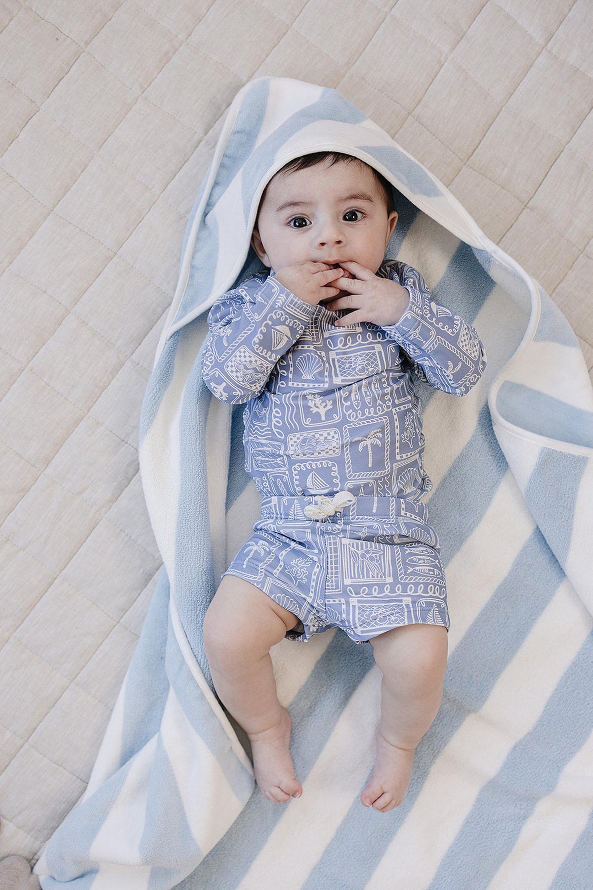 A baby lies on a soft, beige blanket, wrapped in a light blue and white striped hooded towel.