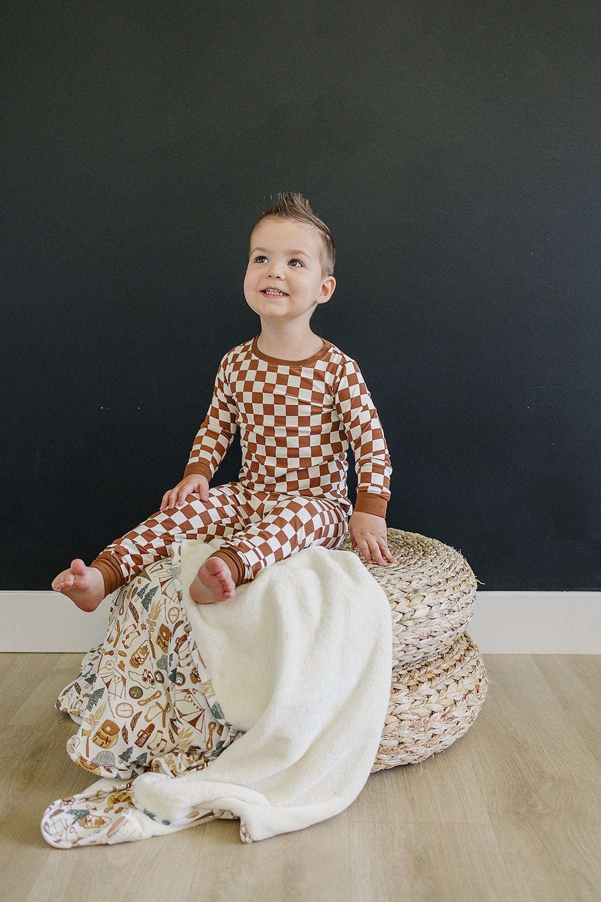 A child in a rust checkered pajama set sits on woven baskets with a cozy blanket draped nearby.