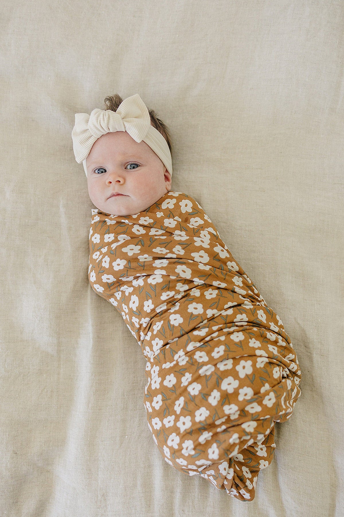 A baby is wrapped in a mustard floral swaddle blanket with a cream headband, lying on a beige surface.