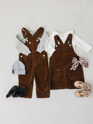 A pair of brown corduroy pinafore dresses for kids is displayed alongside a grey long-sleeve shirt, a beanie, and two pairs of shoes, showcasing a cozy, stylish outfit for children.