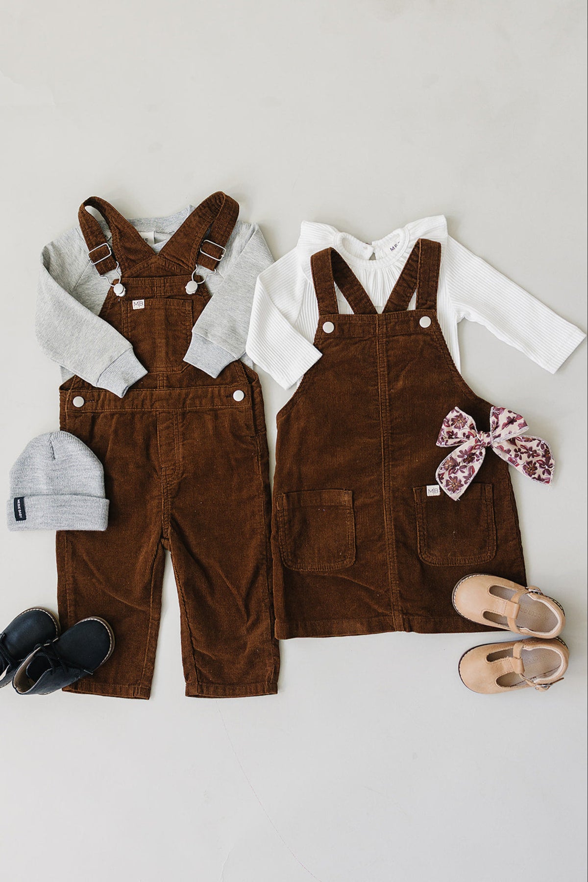 Two matching brown corduroy overalls are displayed alongside a light gray long-sleeve shirt, a gray beanie, and various shoes. The set is perfect for children, combining style and comfort.