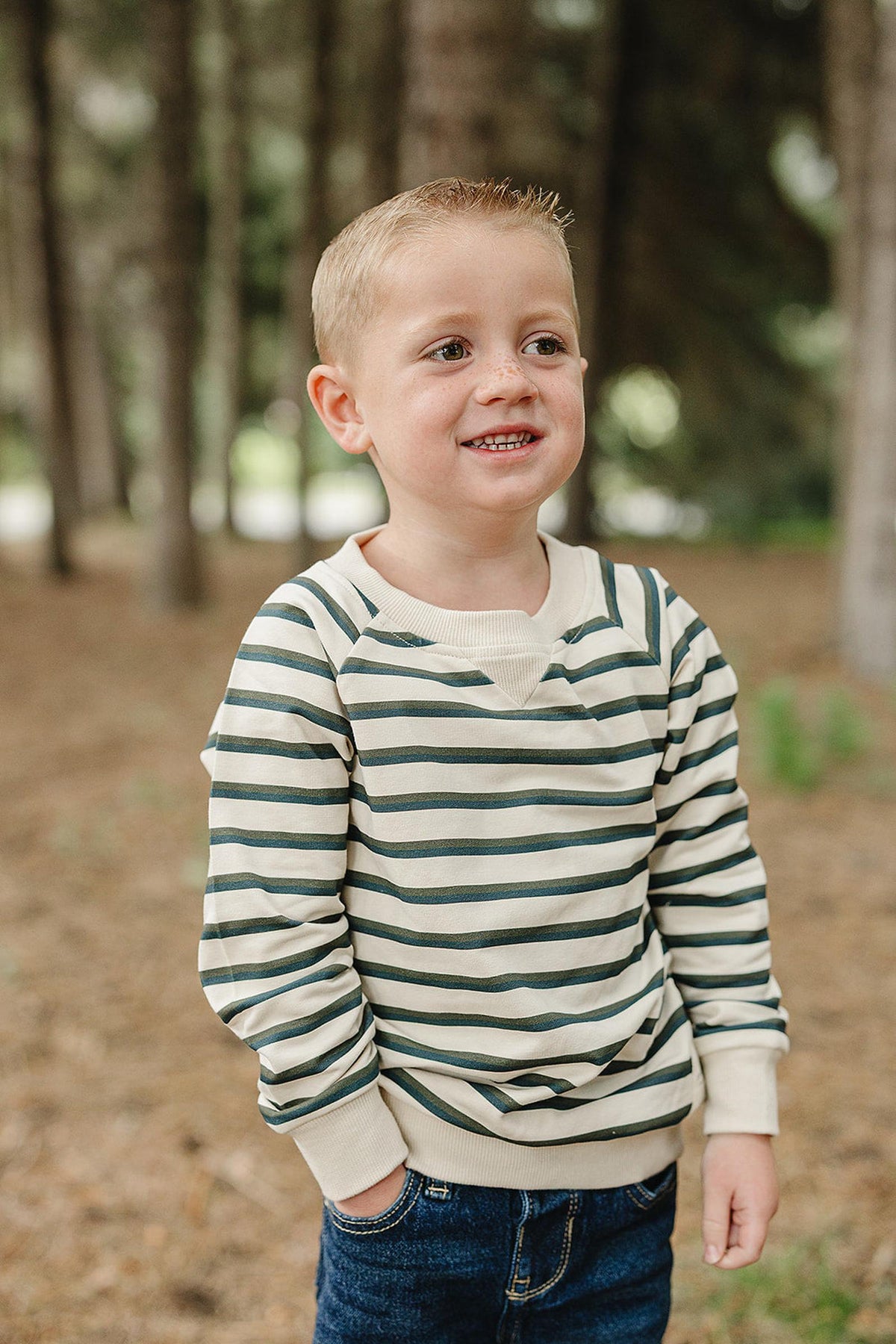 A young boy stands in a wooded area, wearing a soft, striped long-sleeve crewneck t-shirt in green and cream.