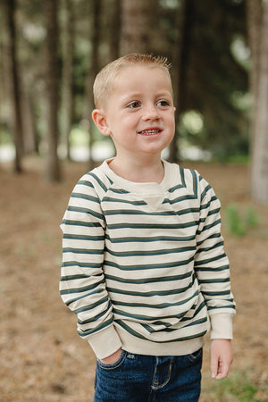 A young boy stands in a wooded area, wearing a soft, striped long-sleeve crewneck t-shirt in green and cream.