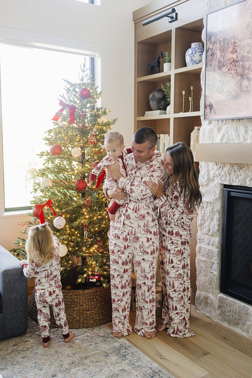 A family in matching holiday-themed bamboo pajama sets poses near a decorated Christmas tree, showcasing a cozy and festive atmosphere. The pajamas feature a classic toile print in red.