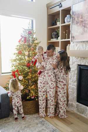 A family in matching holiday-themed bamboo pajama sets poses near a decorated Christmas tree, showcasing a cozy and festive atmosphere. The pajamas feature a classic toile print in red.