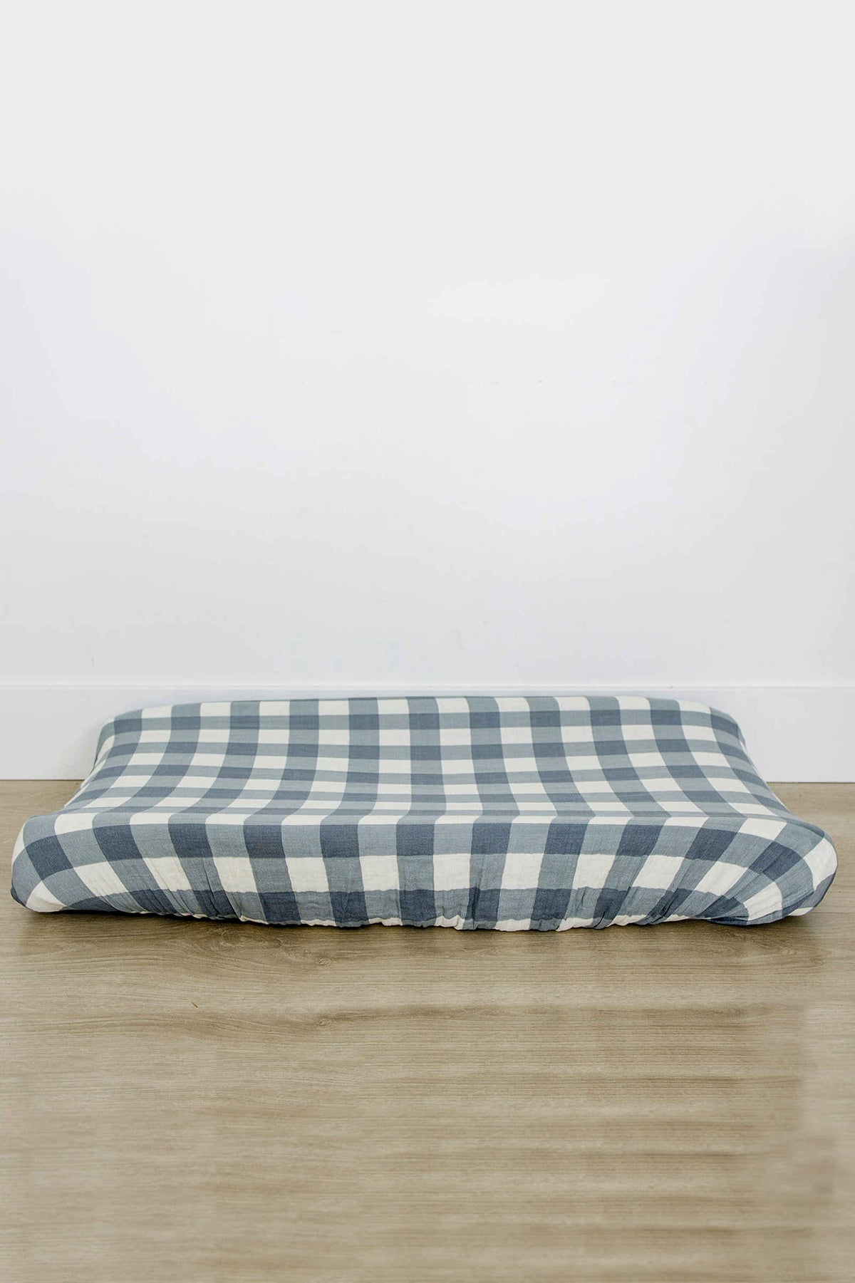 Dusty blue gingham changing pad cover on wooden surface, featuring checkered pattern and elasticized edges for a snug fit.