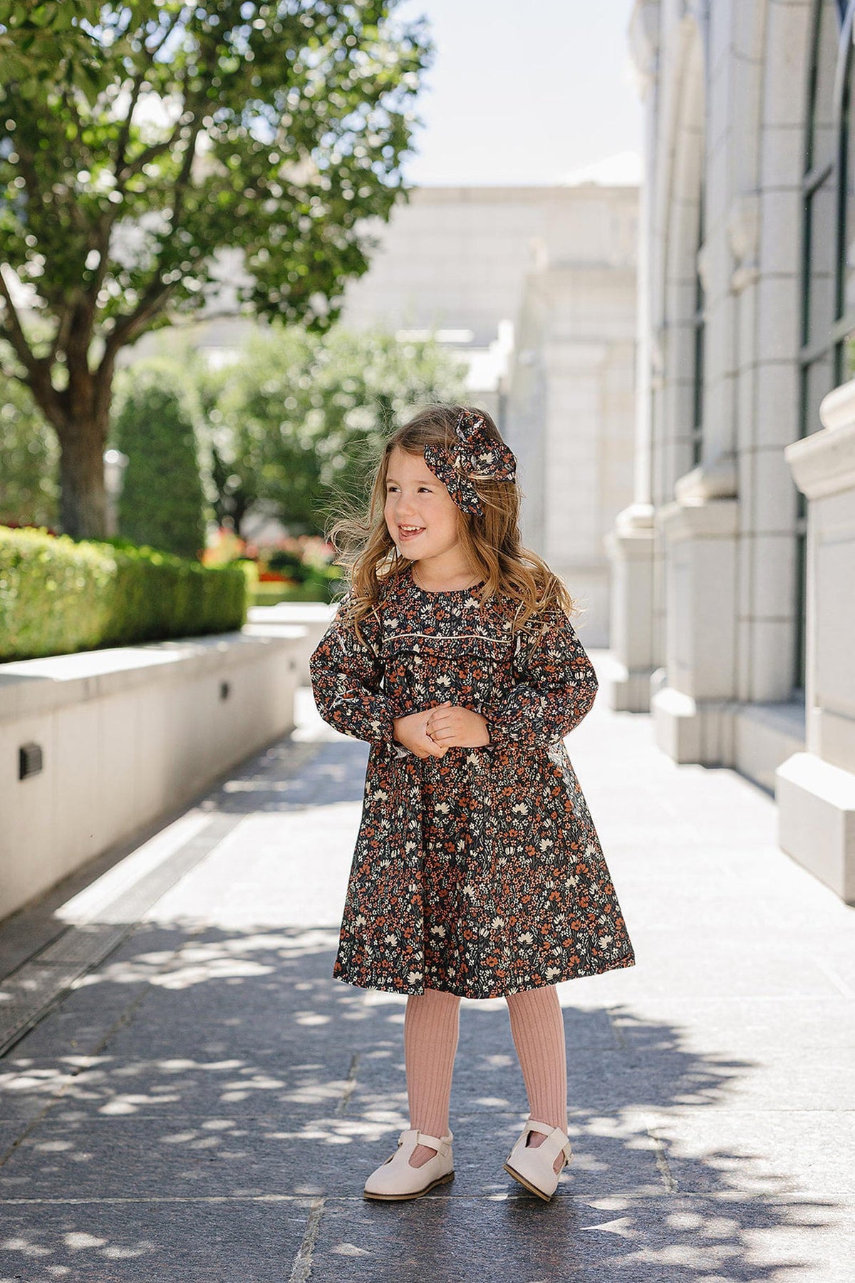 A young girl wearing a floral dress and matching bow stands on a sunlit stone path near a building.