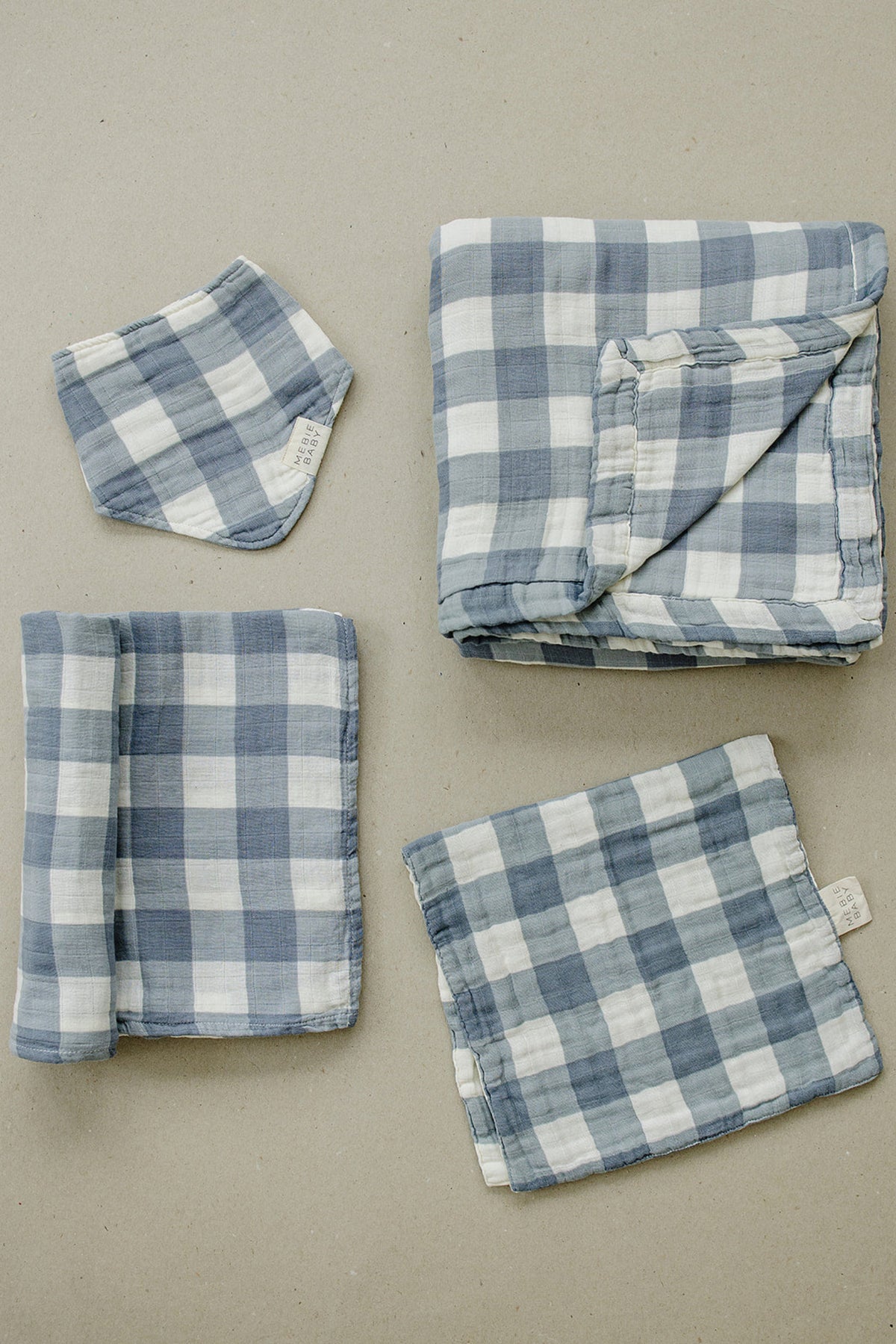 Dusty blue gingham muslin swaddle blankets and accessories laid out on a neutral background, showcasing soft cotton fabric.
