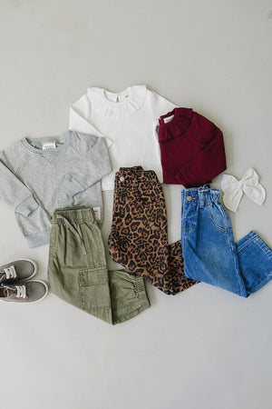A flat lay displays various kids clothing items, including a burgundy ribbed long sleeve top, gray sweatshirt, and patterned leopard pants.