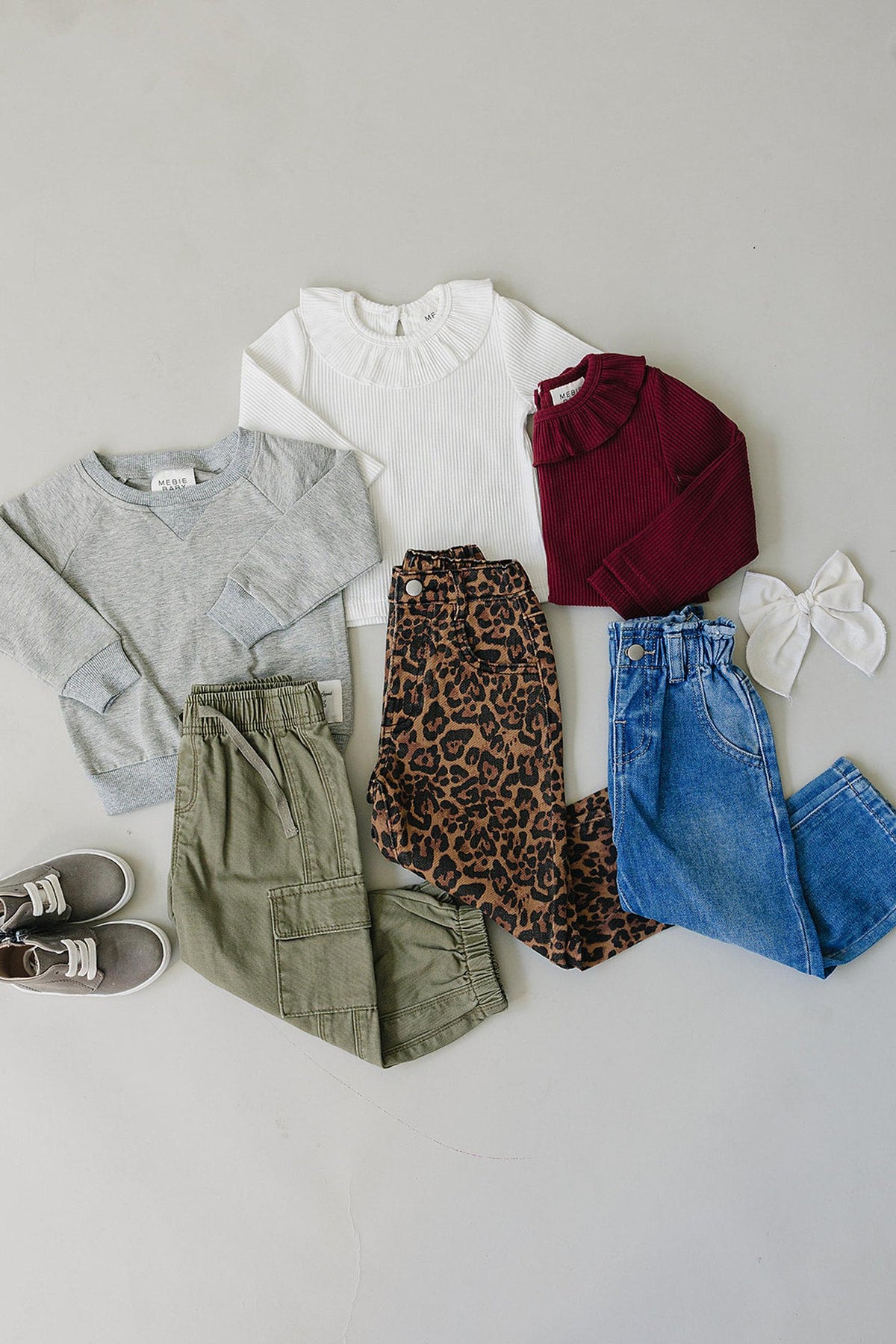 A flat lay showcases a variety of children’s clothing, including a gray sweater, white ribbed top, maroon shirt, leopard print jeans, olive cargo pants, and blue jeans.