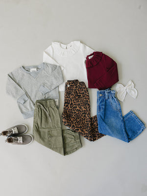 A flat lay displays a variety of childrens clothing, including a gray sweater, white top, burgundy turtleneck, leopard-print pants, and denim jeans, along with a hair bow and sneakers.