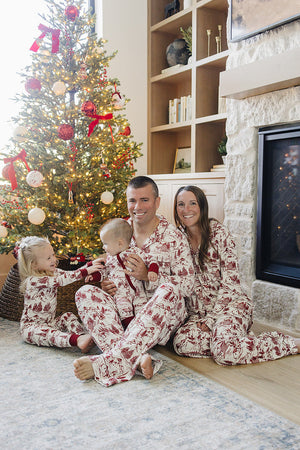 A family sits by a beautifully decorated Christmas tree, all wearing matching red and white holiday toile bamboo pajama sets. The cozy setting features a fireplace and festive decorations.