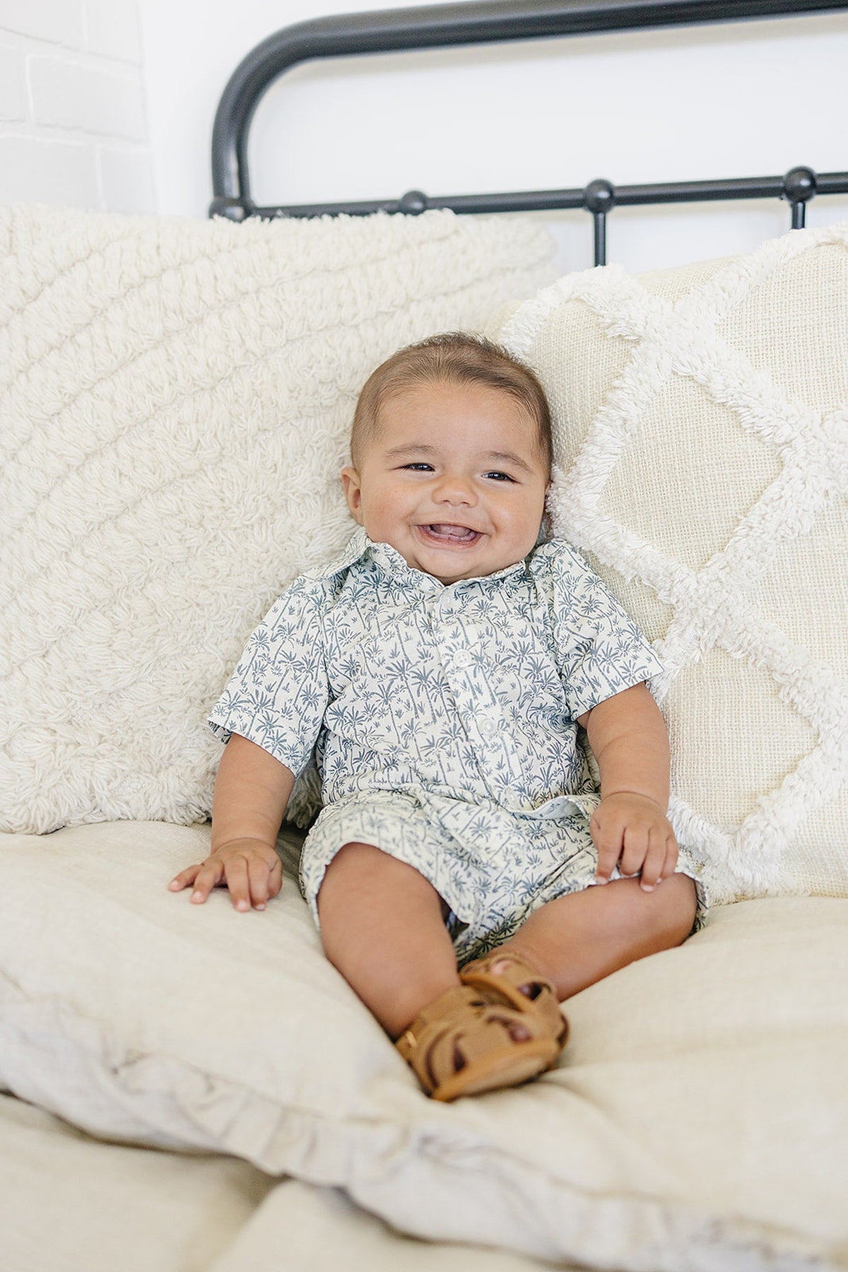 A smiling baby wears a palm tree patterned linen button-up shirt, sitting on a cozy, textured sofa.
