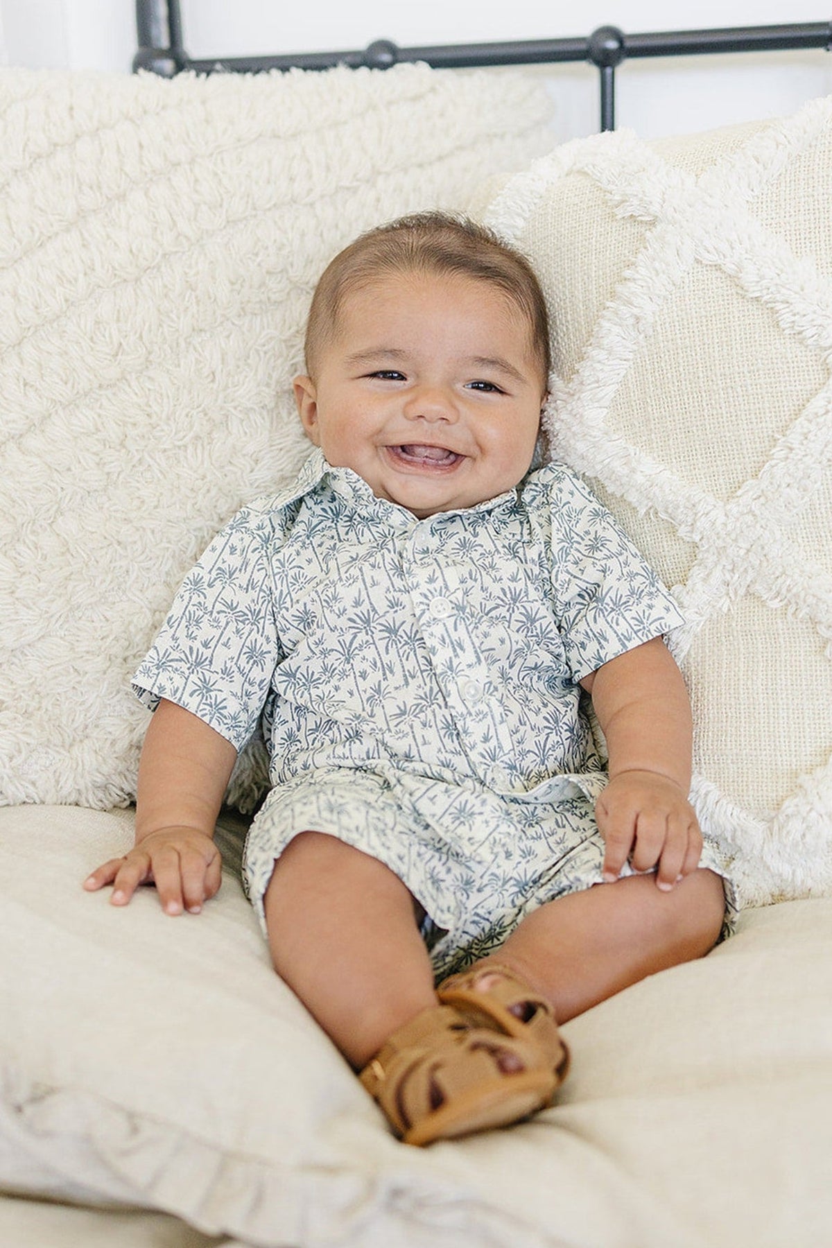 A smiling baby wearing palm tree print shorts and shirt, sitting on a cream-colored, textured sofa.