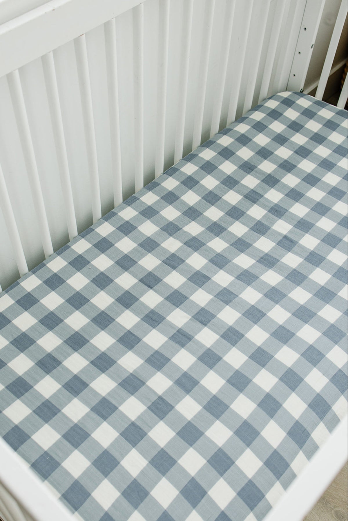 White crib with a dusty blue gingham muslin crib sheet, featuring a classic checkered pattern.