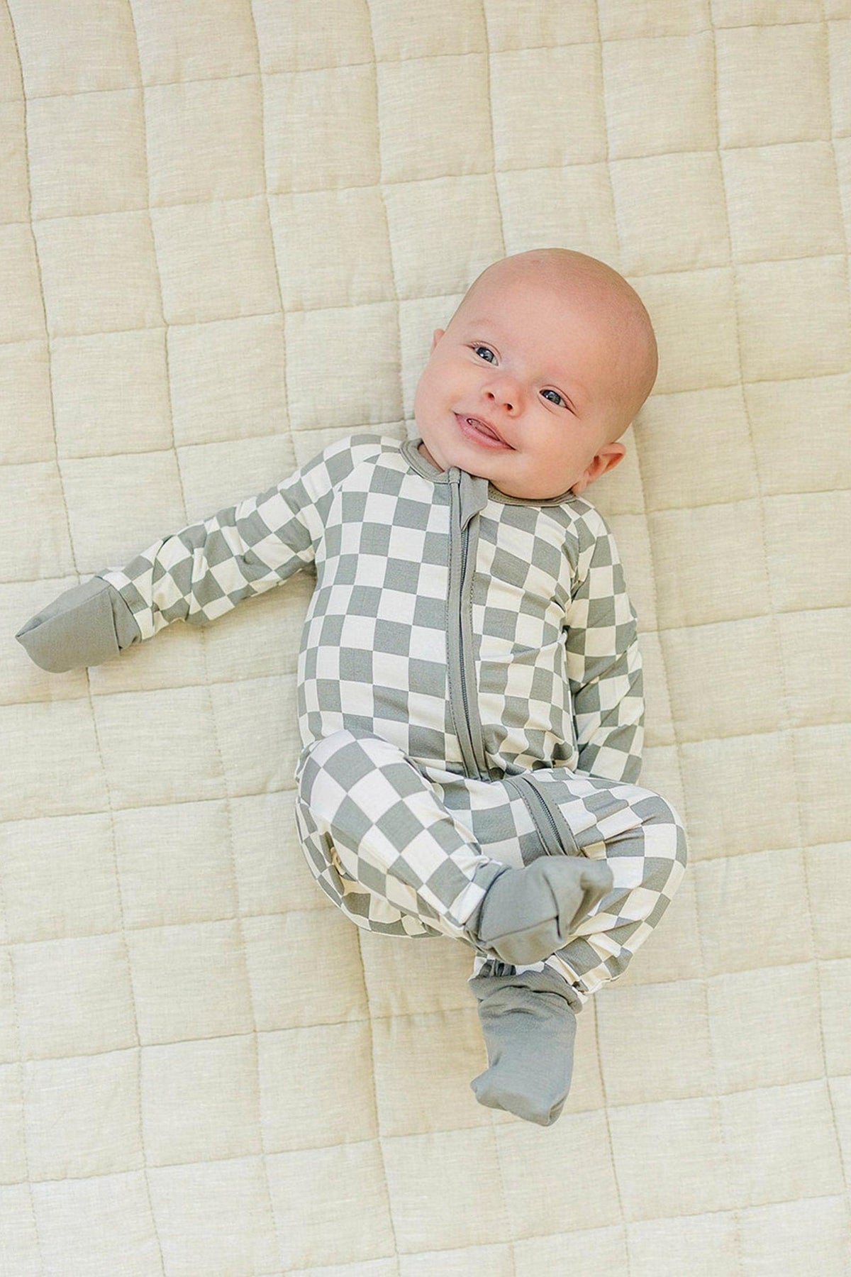 A baby wearing light green checkered zipper pajamas lies on a quilted beige blanket, smiling.