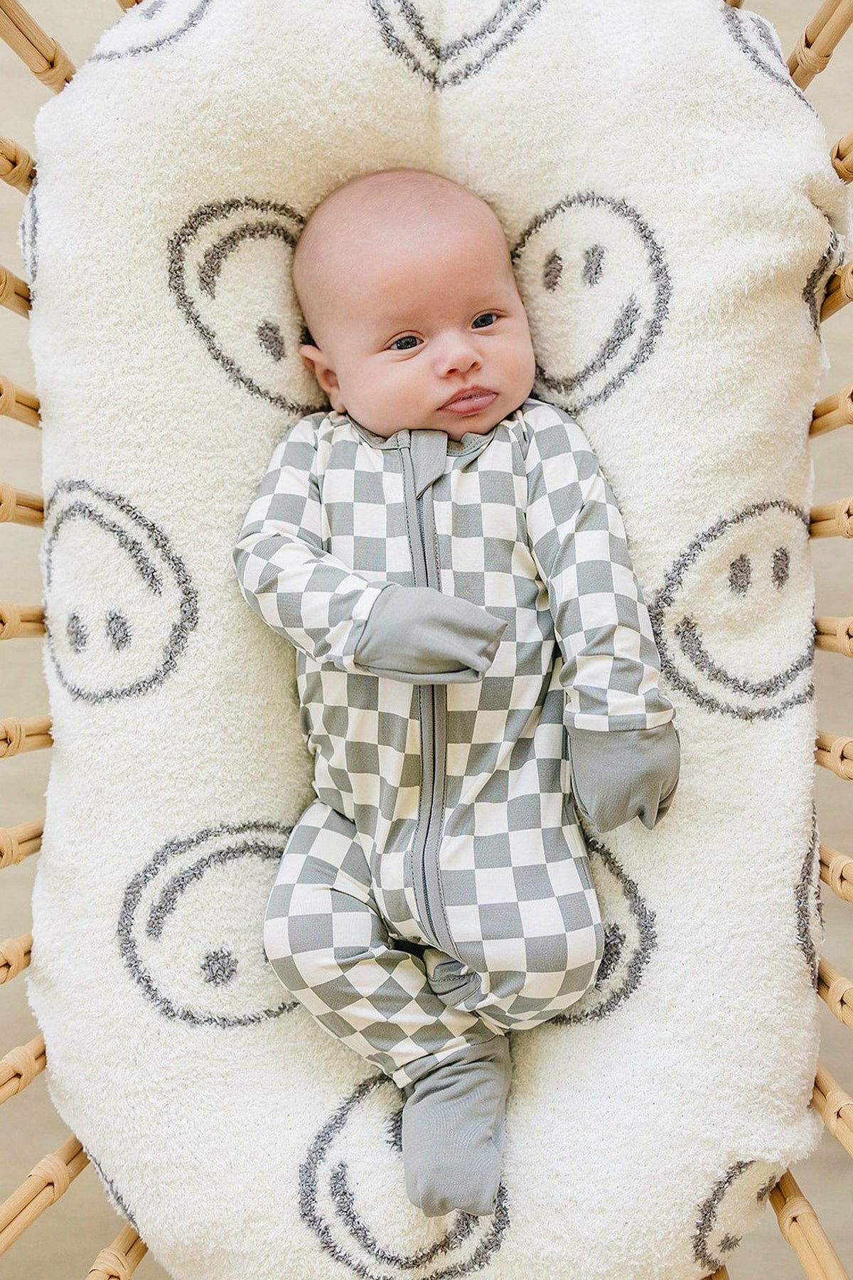 A baby wearing light green checkered pajamas with a zipper, lying on a smiley face-patterned cushion.