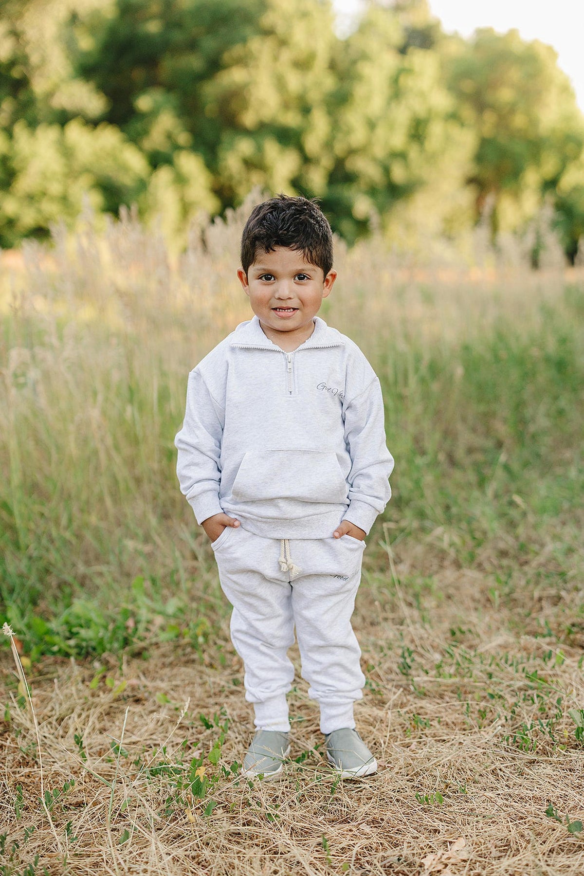 A child in a grey quarter zip sweatshirt set stands in a grassy outdoor setting, smiling.