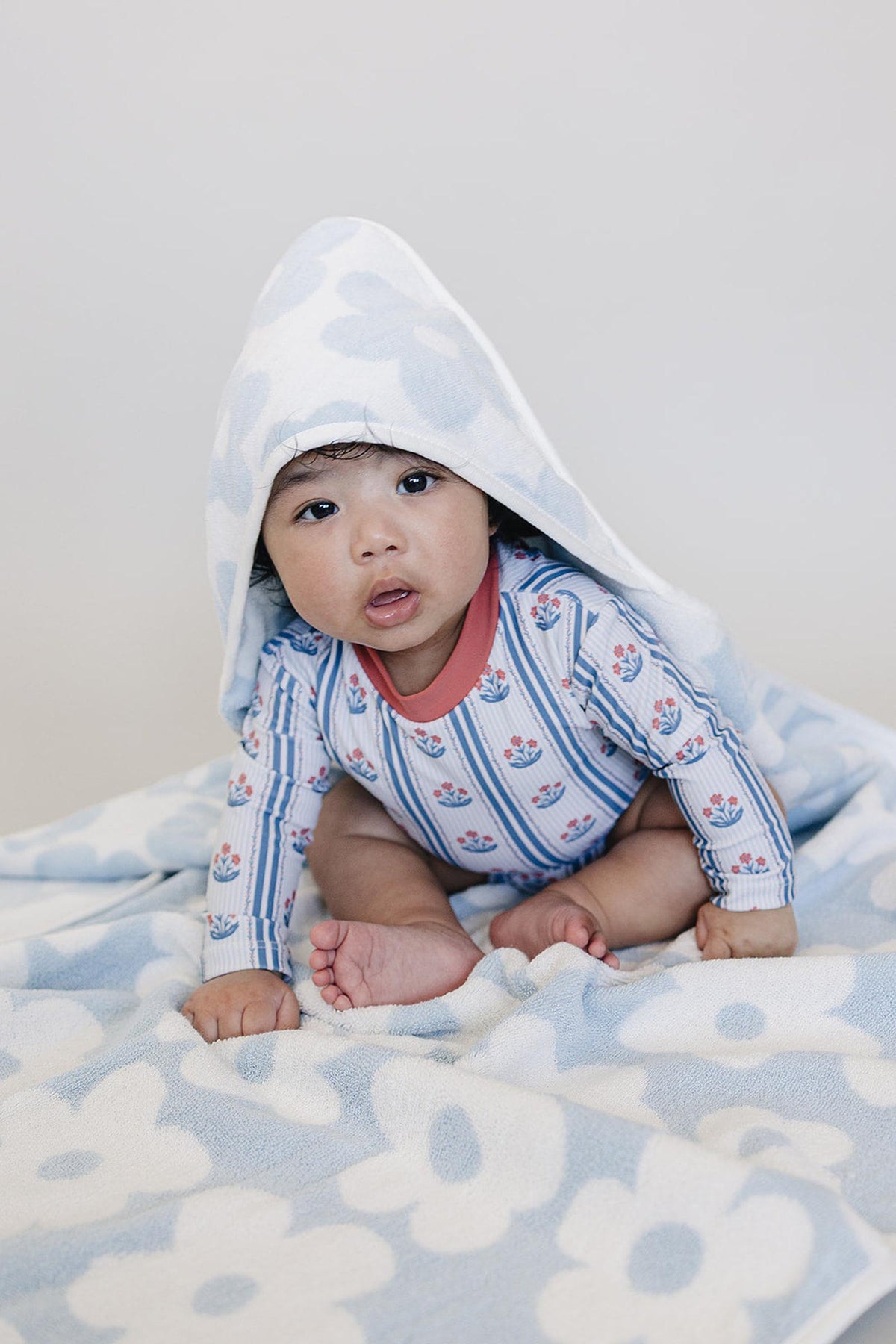 A baby sits on a light blue floral towel, wearing a long-sleeve swimsuit with pink and blue patterns. The hooded towel adds warmth and comfort, perfect for post-bath relaxation.