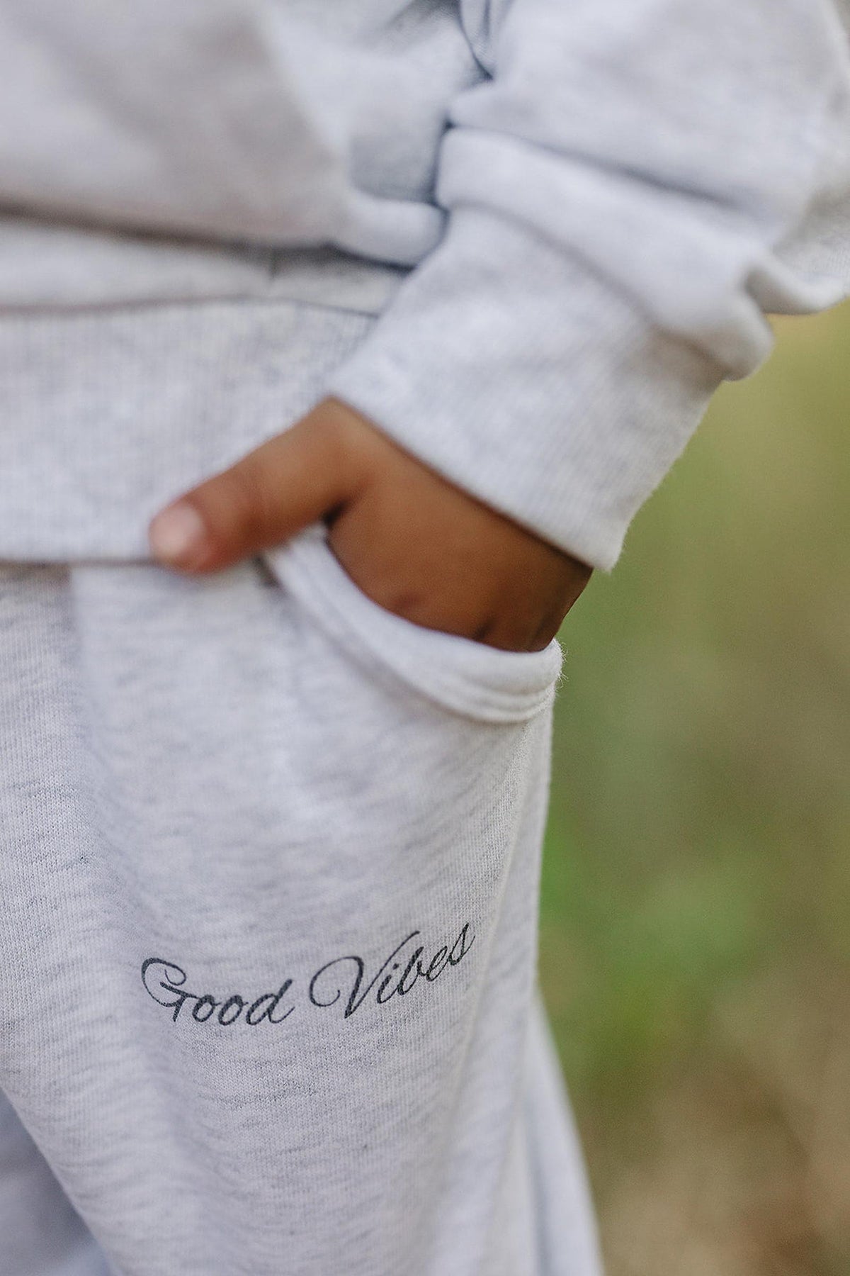 Close-up of a child wearing a grey sweatshirt with Good Vibes text, hand in pocket outdoors.