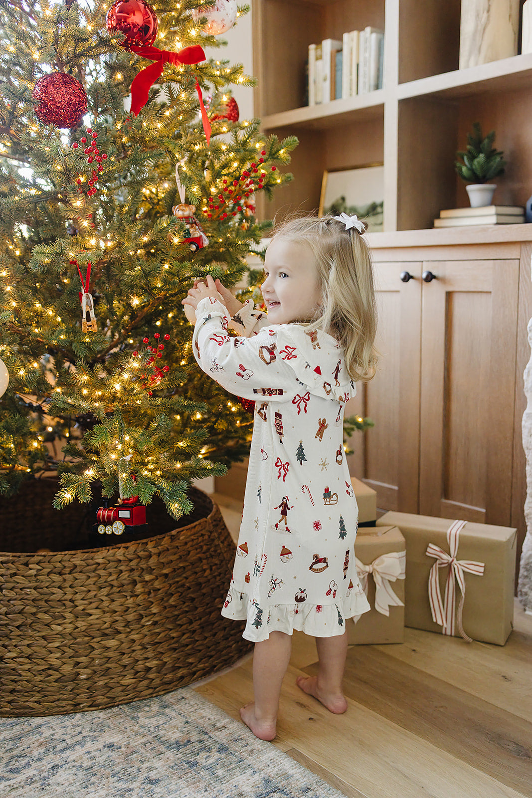 A young girl in a festive, cream-colored gown decorated with holiday patterns stands by a beautifully lit Christmas tree, surrounded by wrapped gifts and a cozy, inviting setting.