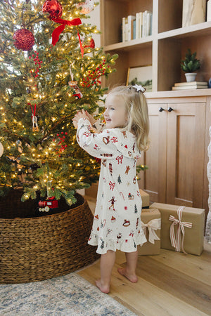 A young girl in a festive, cream-colored gown decorated with holiday patterns stands by a beautifully lit Christmas tree, surrounded by wrapped gifts and a cozy, inviting setting.