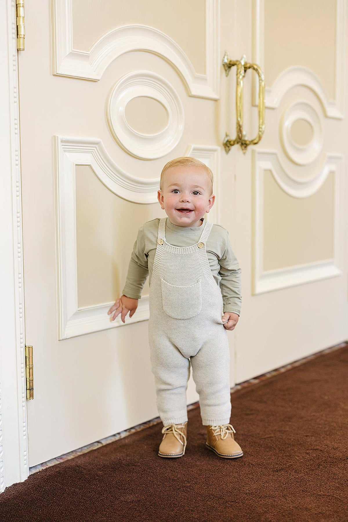 A smiling baby in grey knit overalls and tan shoes stands in front of ornate cream doors.
