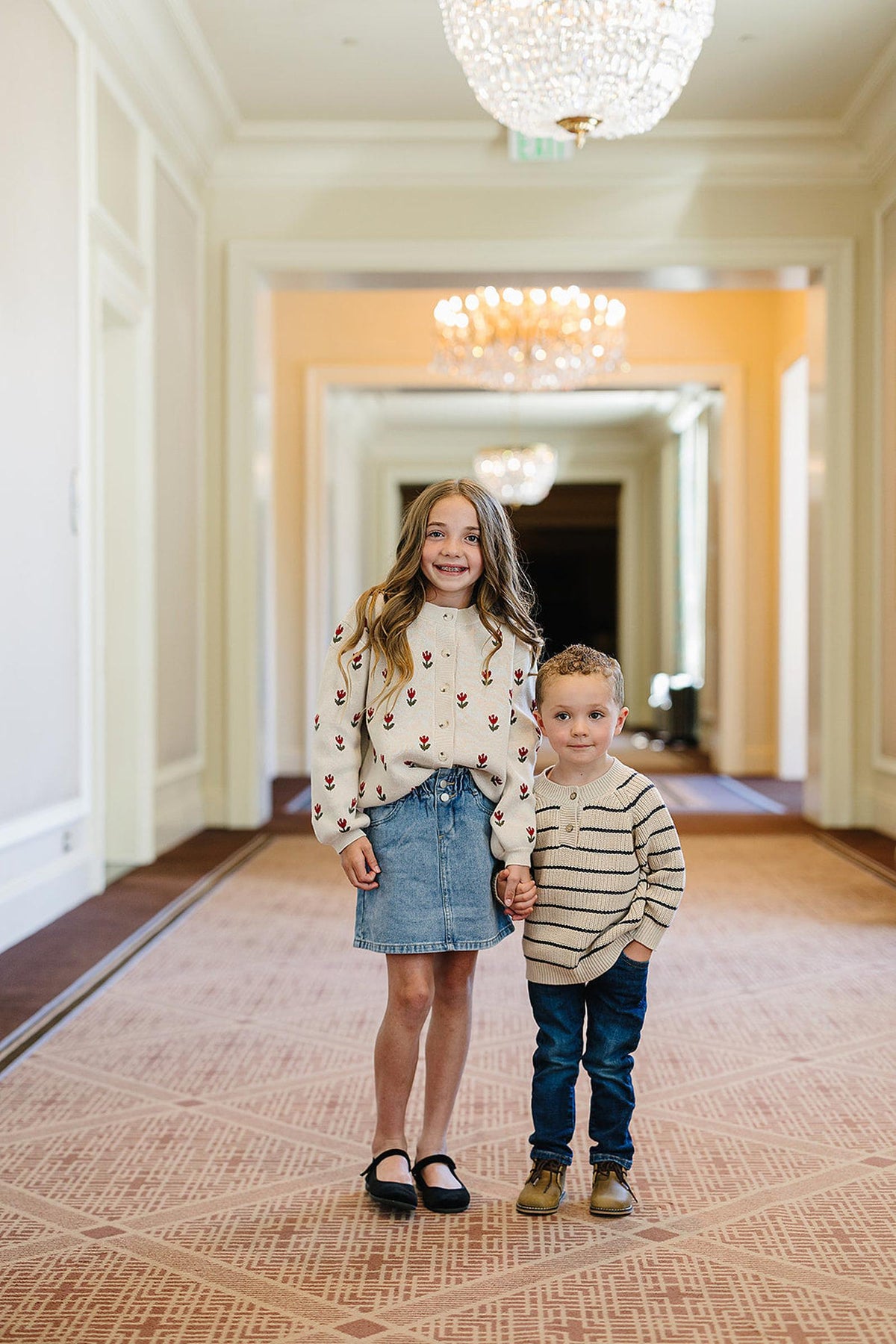 Two children in a hallway, one in a floral sweater and denim skirt, the other in a striped sweater.