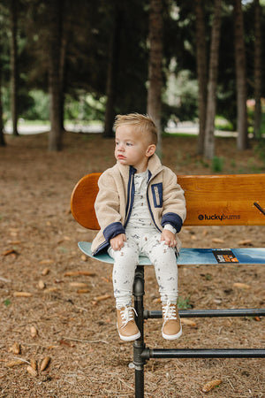 A toddler sits on a wooden bench in a forest, wearing a cozy beige jacket and ski-print pajamas with brown.