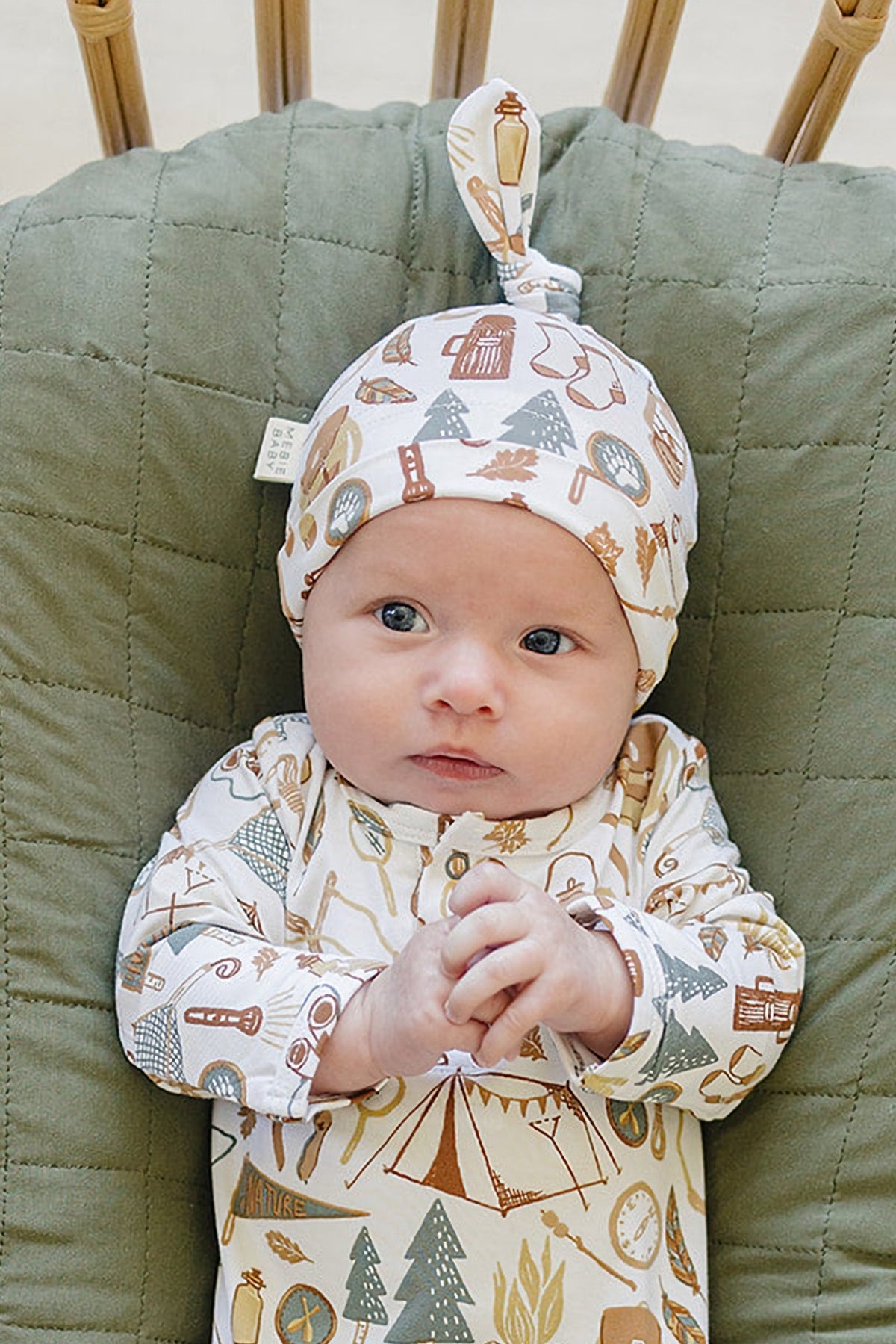 A newborn baby wears a patterned knot hat and matching outfit featuring camping-themed illustrations, resting on a quilted green blanket.