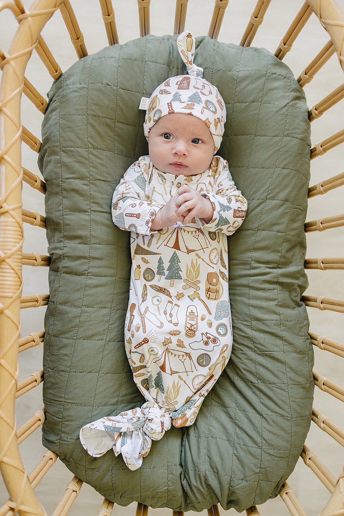 A newborn lies on a green quilted pad, wearing a patterned bamboo gown and matching knot hat featuring camping-themed designs.