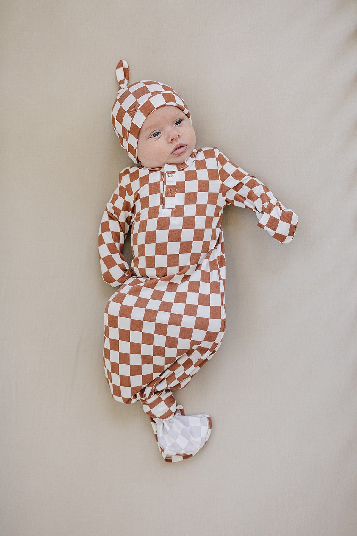 A baby lies on a beige surface, wearing a rust checkered gown and matching hat, showcasing soft bamboo fabric.